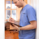 Ein Arzt im Arztkittel schreibt auf einem Klemmbrett in einem Flur mit Holzboden und einem Fenster
