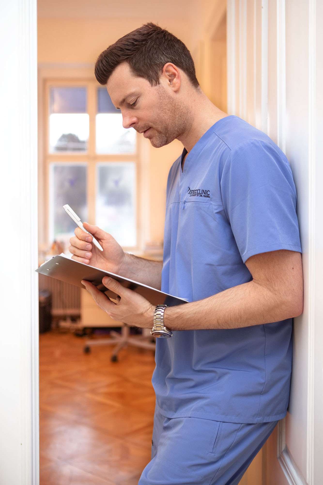 Ein Arzt im Arztkittel schreibt auf einem Klemmbrett in einem Flur mit Holzboden und einem Fenster