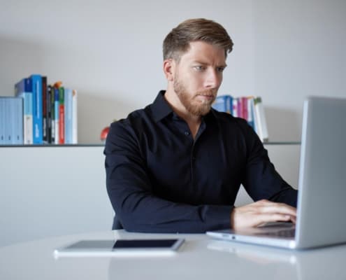 Ein Mann sitzt an einem Tisch mit einem Laptop vor ihm und einem Bücherregal hinter ihm