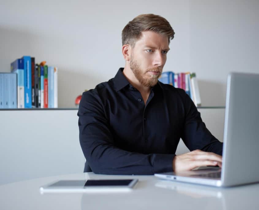Ein Mann sitzt an einem Tisch mit einem Laptop vor ihm und einem Bücherregal hinter ihm