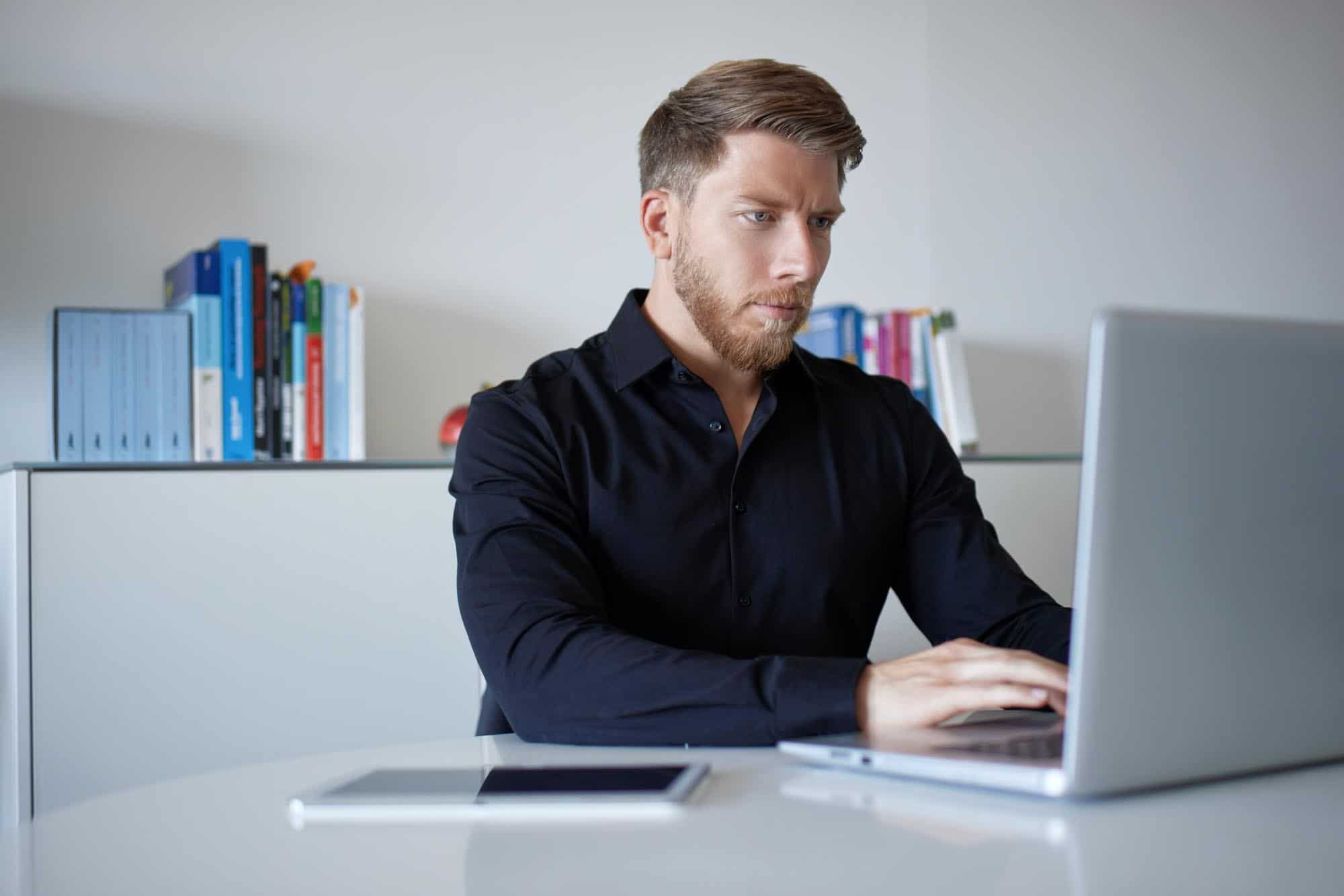 Ein Mann sitzt an einem Tisch mit einem Laptop vor ihm und einem Bücherregal hinter ihm