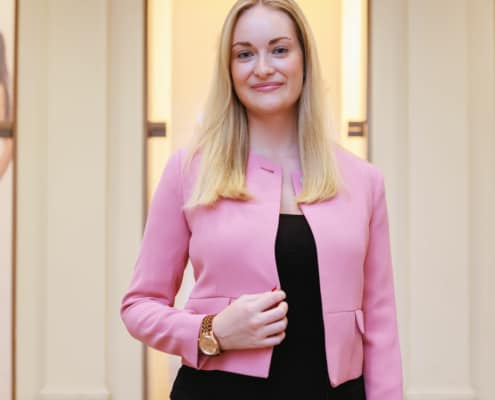 Eine Frau in einer rosa Jacke und einer schwarzen Hose, die vor einem Gebäude steht.