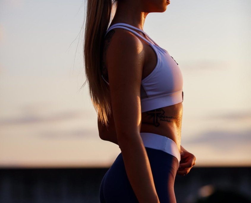 Eine Frau in einem weißen Oberteil und blauen Leggings steht vor einem Sonnenuntergang