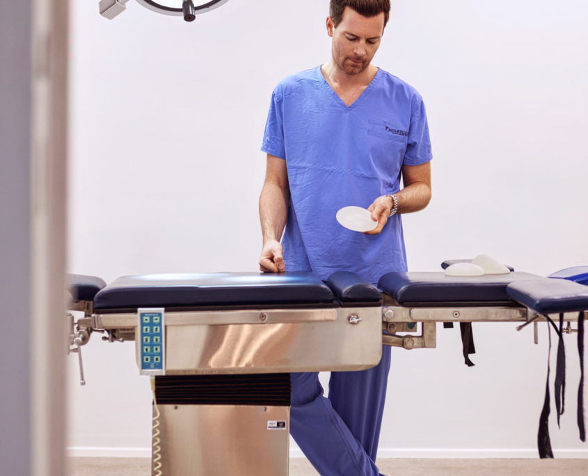 ein arzt im arztkittel steht an einem operationstisch mit einem brustimplantat in der hand und einer lichtlampe über ihm
