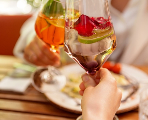 eine Frau stoßt mit einem Weinglas mit verschiedenen Früchten darin gegen ein anderes Weinglas mit verschiedenen Früchten darin
