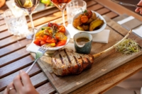 Ein Teller mit einem Tomahawk Steak und gegrillten Gemüse auf einem Tisch mit Weingläsern und einer Frau, die eine Gabel hält