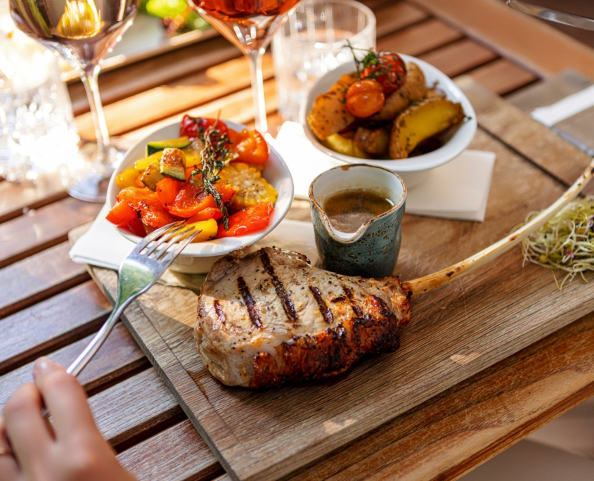 Ein Teller mit einem Tomahawk Steak und gegrillten Gemüse auf einem Tisch mit Weingläsern und einer Frau, die eine Gabel hält