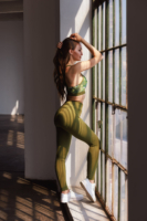 Ein weibliches Fitness Model in grünen Pairadize Fitness Outfit steht an einem Fenster und schaut hinaus.