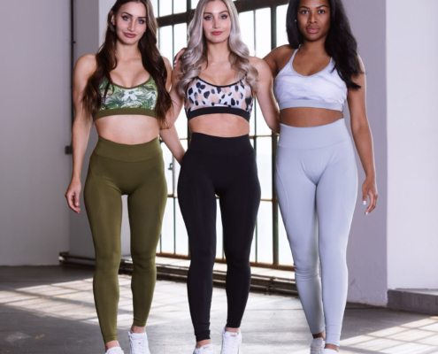 Drei weibliche Fitness Models posieren in verschieden farbigen Fitnessoutfits in einem Raum mit großen Fenstern