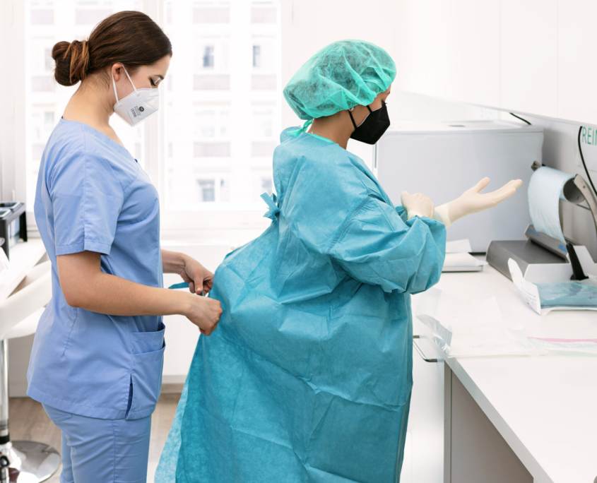Eine Krankenschwester bindet den Kittel einer Ärztin zu die sich die Hände wäscht und auf eine OP vorbereitet