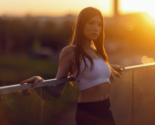 Ein weibliches Fitness Model mit weißen Sport BH und dunkler Leggins lehnt bei untergehender Sonne an eine Glaswand